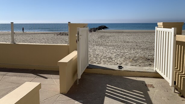 Vista de una playa con entrada protegida, un camino de hormigón y una persona caminando.