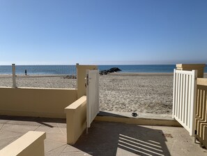 Une vue sur la plage avec une entrée fermée par un portail, un chemin en béton et une personne qui se promène sur la plage.