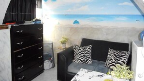 A room with a beach mural, a ceiling fan, a black sofa with zebra-patterned pillows, a small table with a plant, and a black storage unit with drawers. - Blue Horizon Guest House - 3-Bedroom (Warwick)