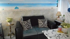 A room with a beach mural on the wall, a black sofa with patterned cushions, a small table with a lamp, and a potted plant. - Blue Horizons Guest House - Room # 7 (Hamilton)