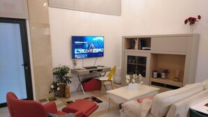 A modern living room with a flat-screen TV mounted on the wall, a red chair, a beige sofa, a small table with a vase of flowers, and a bookshelf. - Beachside apartment (Santander)
