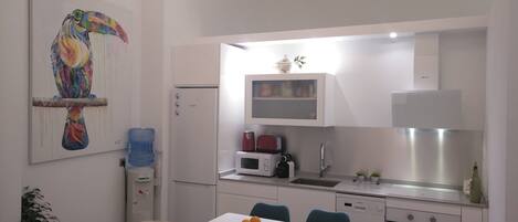 Cocina compacta con refrigerador, microondas y lavavajillas. En primer plano, una mesa de comedor con frutas.