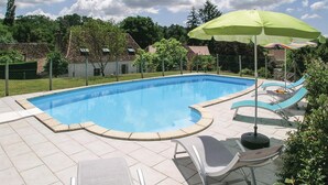 En rektangulär swimmingpool med en böjd kant, omgiven av en terrass med kakelplattor. Där finns solstolar och ett grönt parasoll.