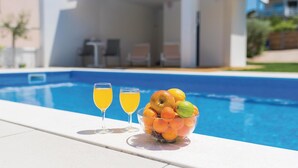 Een zwembadterras met een helderblauw zwembad, twee glaasjes sinaasappelsap en een schaal fruit.