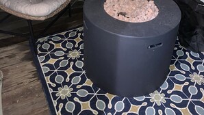 Eine Terrasse mit einem runden Feuerkorb, zwei gepolsterten Stühlen, einem kleinen Tisch und einem gemusterten Teppich.