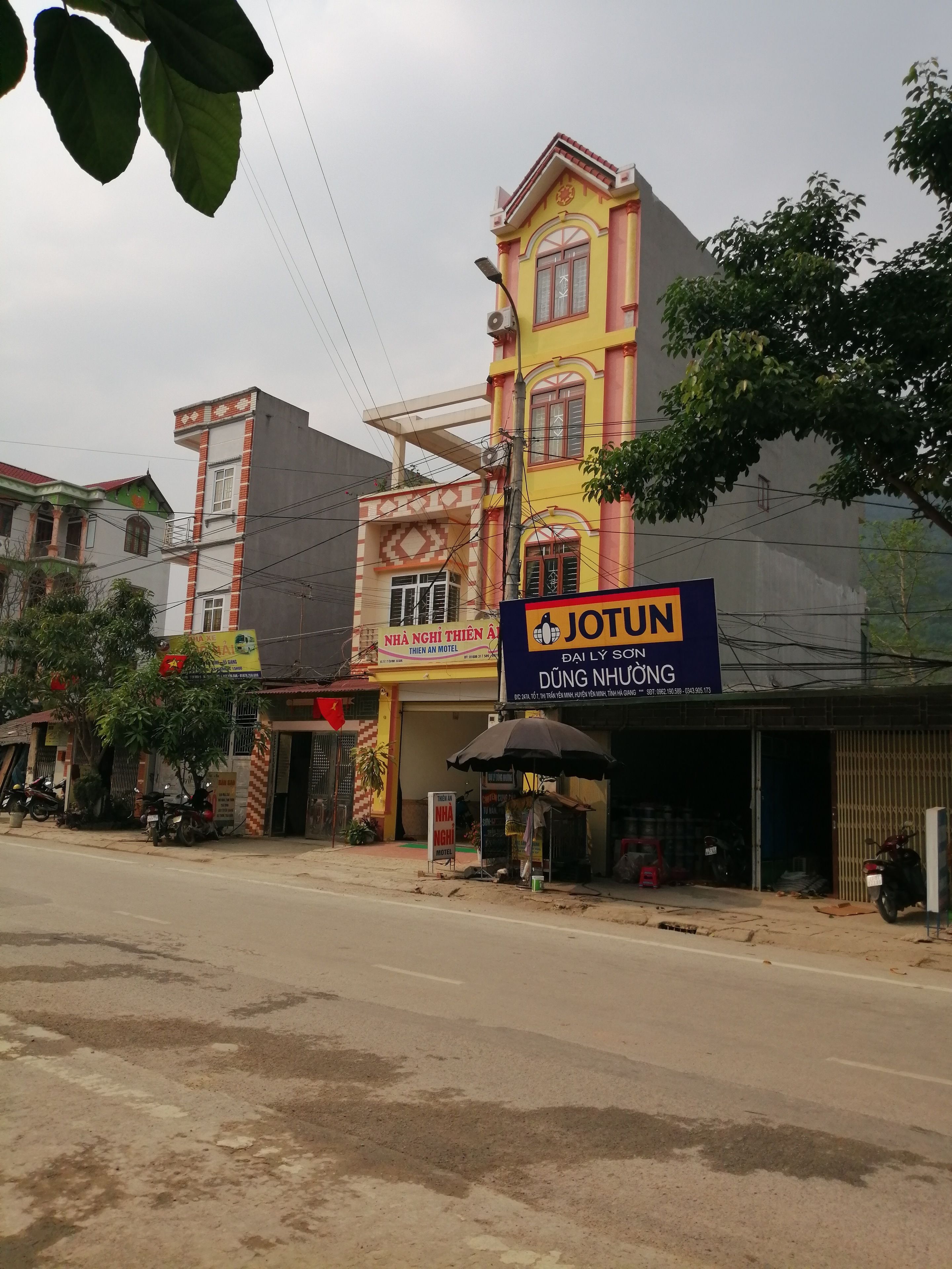 A building with a prominent yellow and red sign for IOTUN, a hotel with a balcony and a red roof, and a small shop with an umbrella.