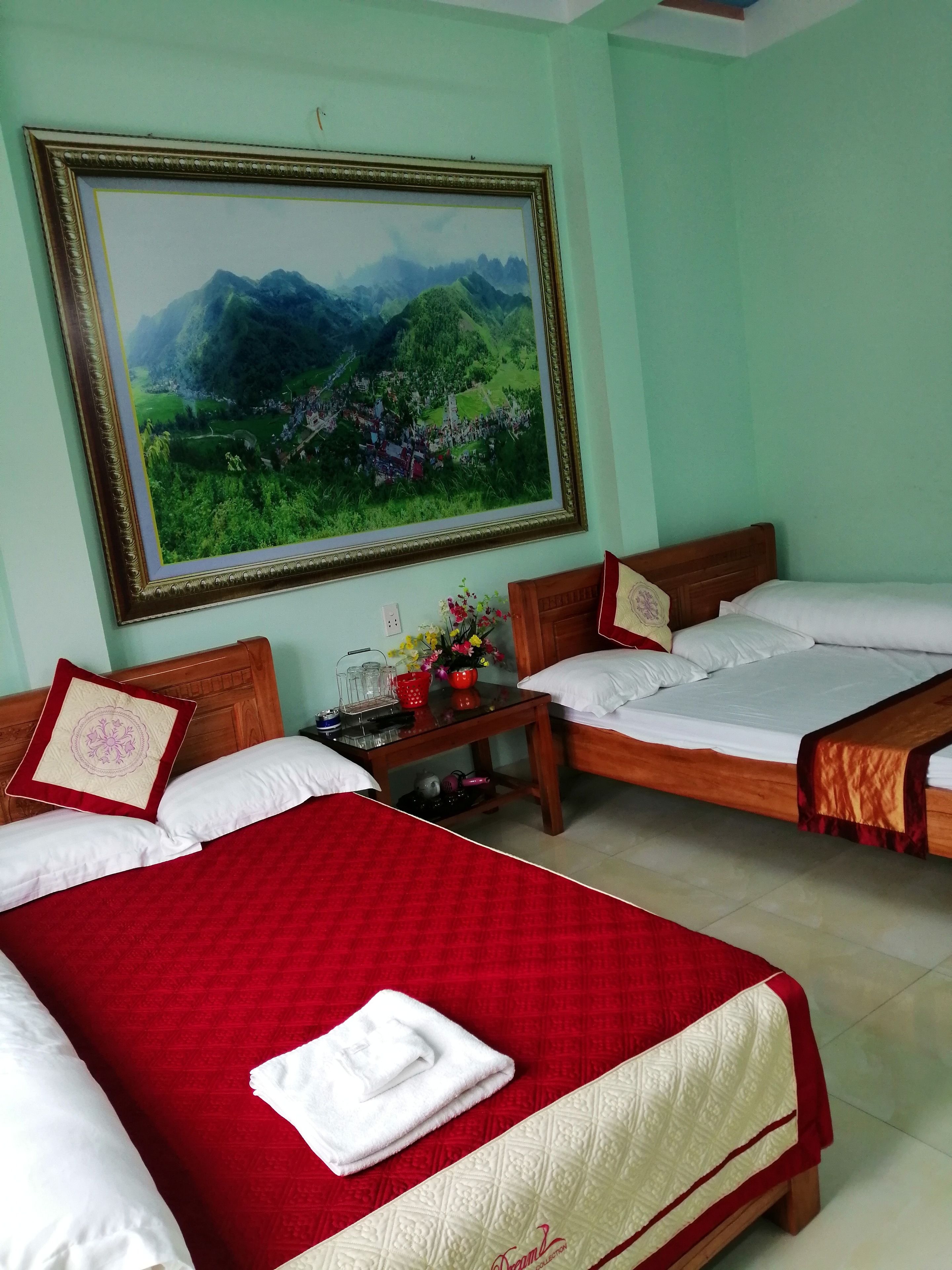 A hotel room with two beds, a large framed landscape painting, and a small table with a vase of flowers.