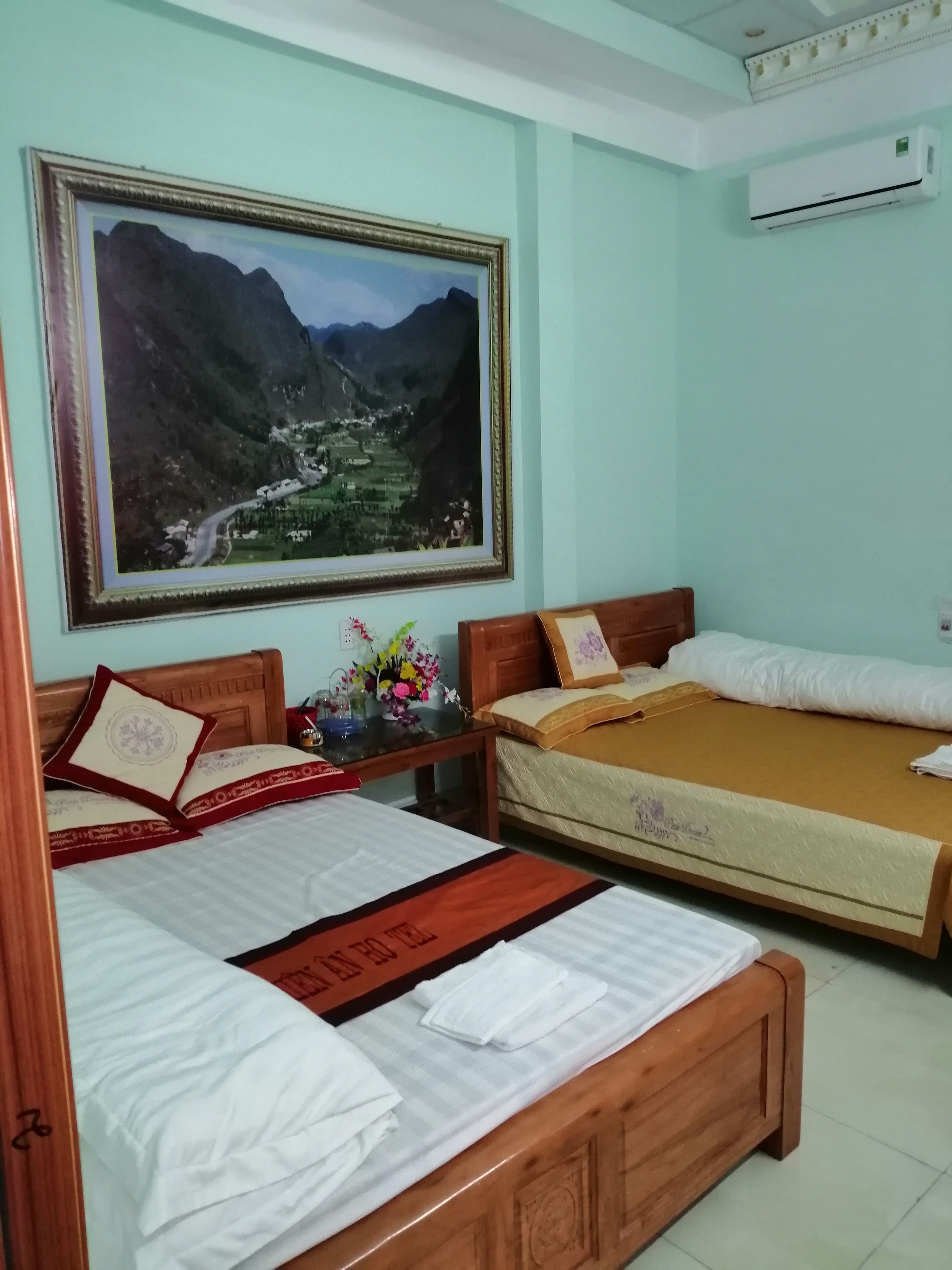 A hotel room with two beds, a large framed landscape painting, a small table with a vase of flowers, and an air conditioning unit.