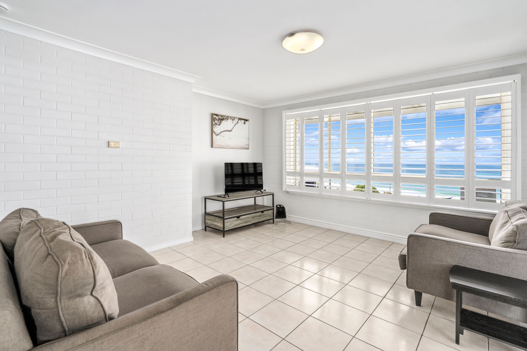 A spacious living room with a large window offering a view of the ocean, a flat-screen TV on a stand, and two sofas.