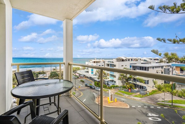 A balcony with a glass table and chairs overlooking a beach and residential buildings.