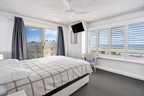 A bedroom with a bed, a television, a window with blinds, and a view of the sea.