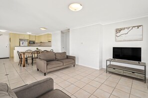 Ein Wohnzimmer mit einer Couch, einem Esstisch mit Stühlen, einem Fernseher auf einem Schrank und einer Küche im Hintergrund.