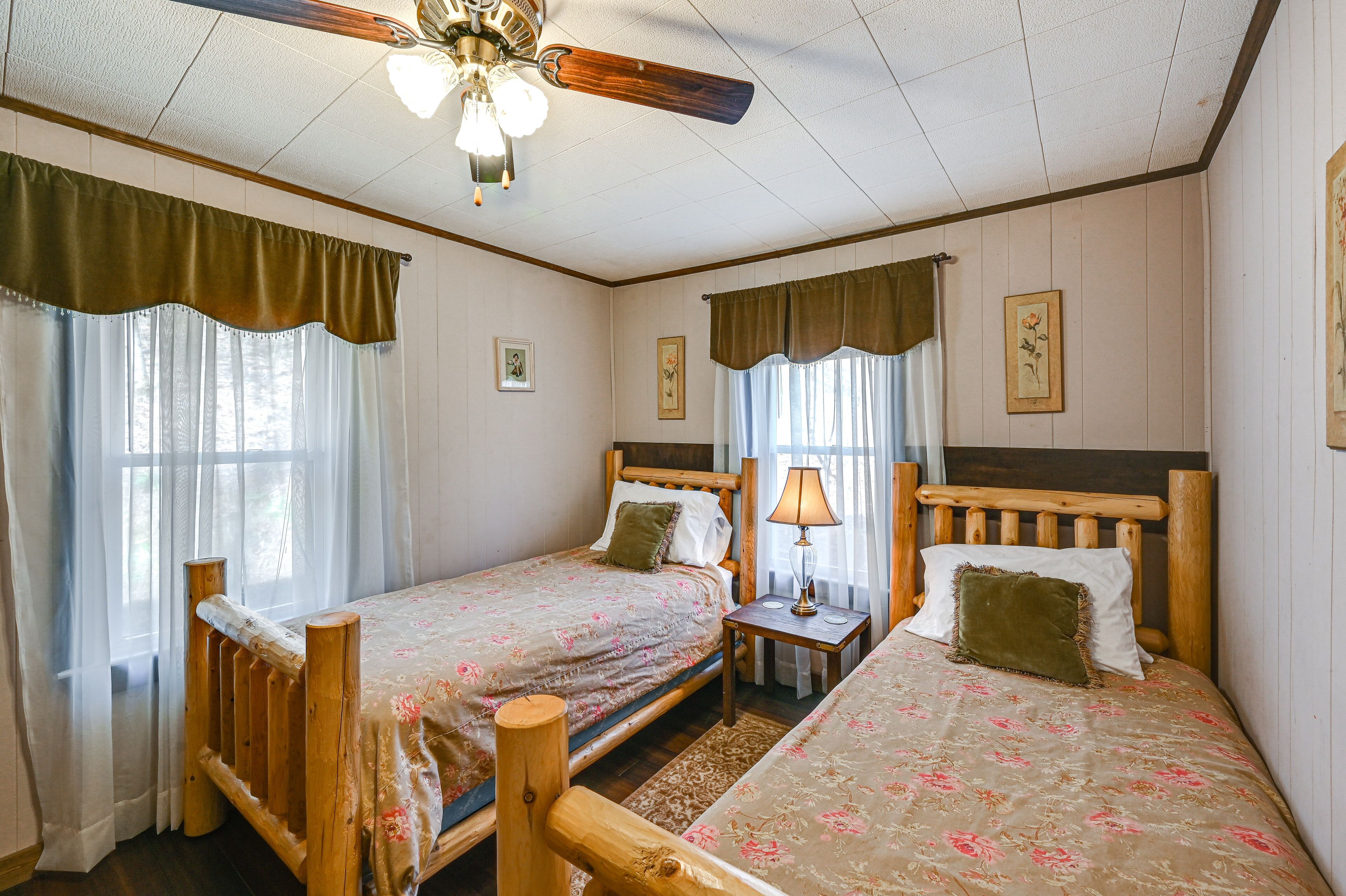 Quarto com duas camas de madeira, cada uma com mesa de cabeceira, abajur e janela com cortinas.