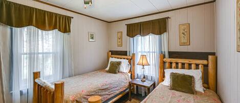 Quarto com duas camas de madeira, cada uma com mesa de cabeceira, abajur e janela com cortinas.