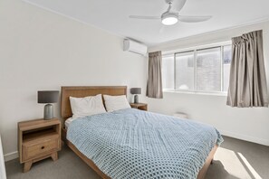 A bedroom with a bed, bedside tables, a ceiling fan, an air conditioning unit, a window with curtains, and a view of the outdoors.
