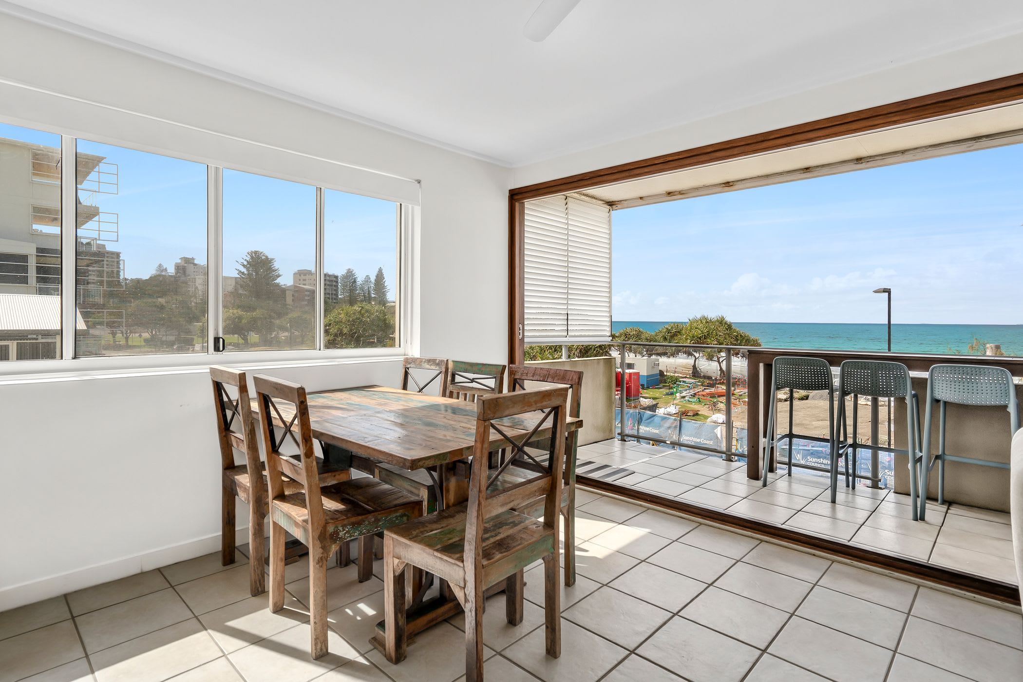 A dining area with a wooden table and chairs, large windows, and a view of the beach.