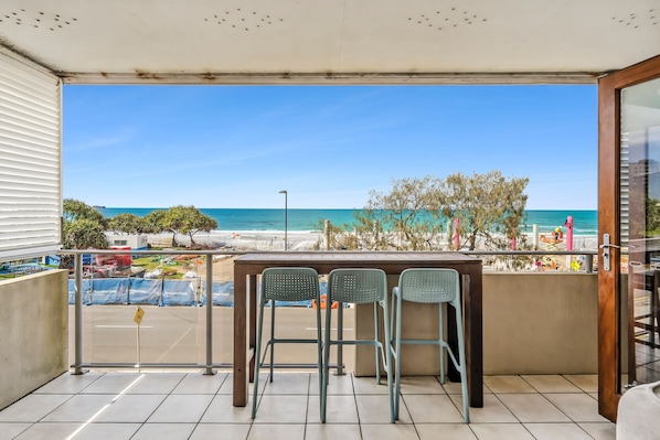 A balcony with a view of the beach, two metal chairs, and a table.