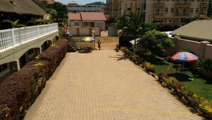 A paved walkway with a white railing on one side, leading to a building with a red roof. There are two umbrellas and a bench in the center. - United Motel Entebbe (Entebbe)