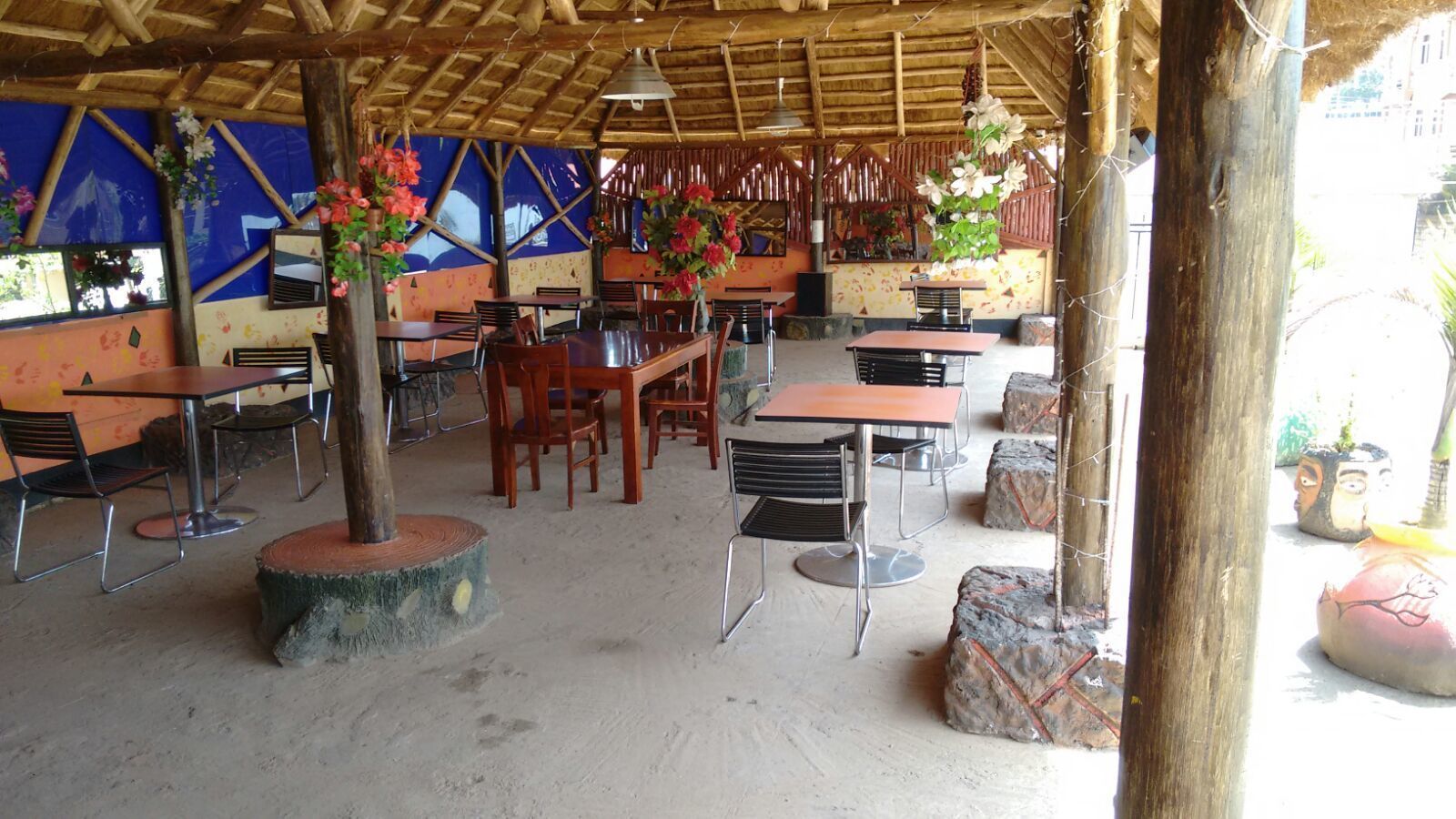 Un espace de restauration en plein air, avec des tables et des chaises, entouré de plantes décoratives et surmonté d’une structure à toit de chaume.