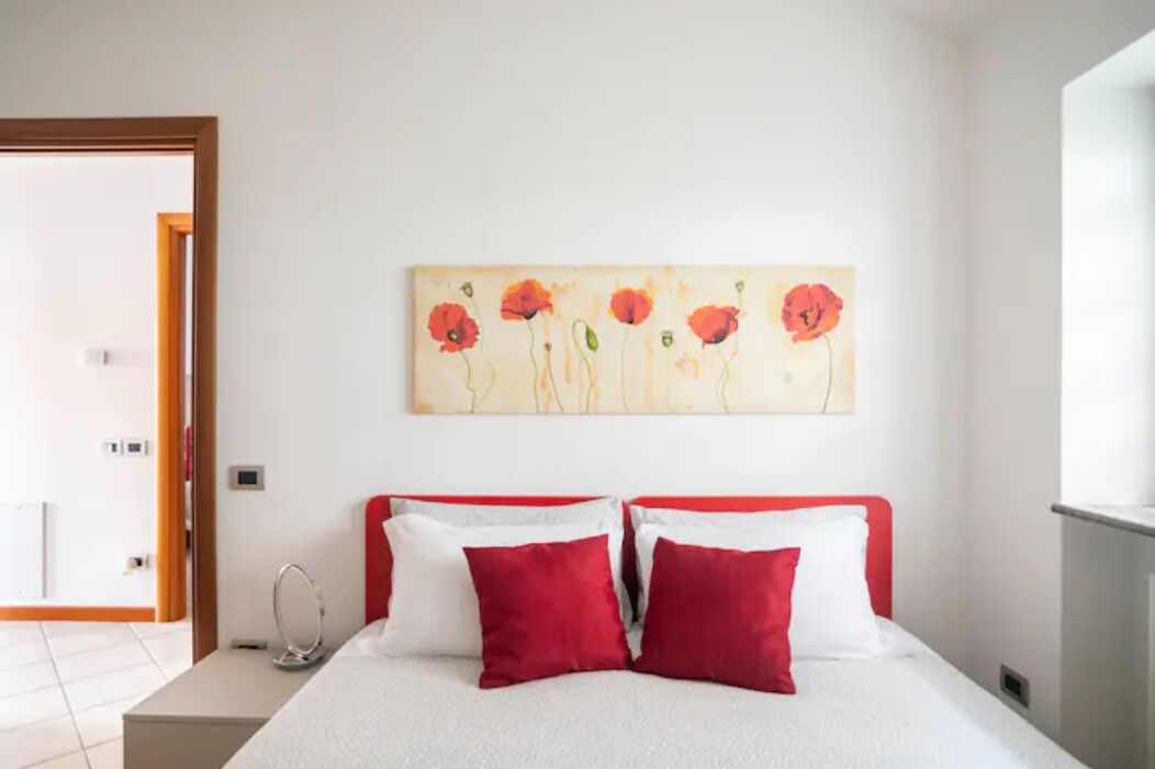 A bedroom with a bed, red and white pillows, a bedside table, and a wall-mounted painting of flowers.