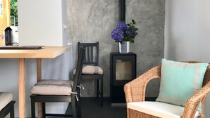 Ein modernes Wohnzimmer mit Esstisch, zwei Stühlen, einem Korbssessel mit Kissen, einer Wanduhr und einem Kamin mit Blumenvase.