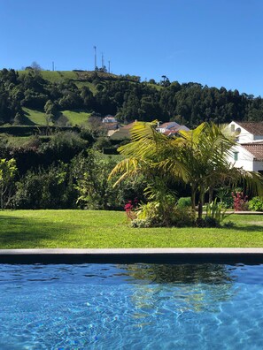 Una piscina con agua cristalina, rodeada de exuberante vegetación y una colina con casas al fondo.