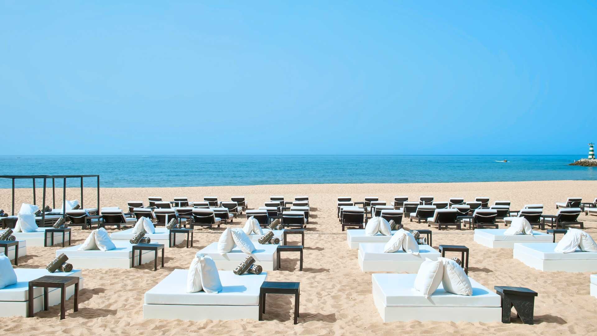 A beachside hotel with rows of white sunbeds and lounge chairs, a clear blue sky, and a calm sea.