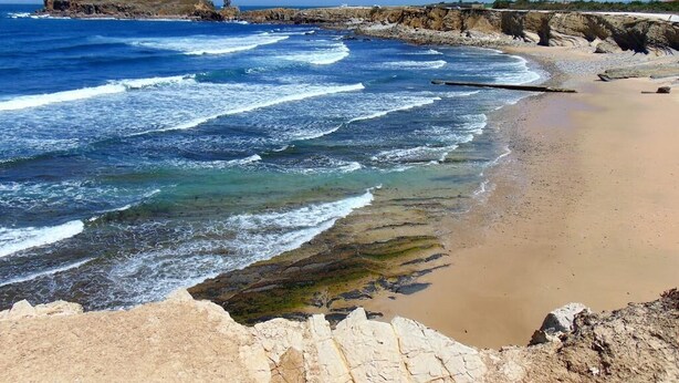 Uma praia costeira com águas azuis cristalinas, areal arenoso e falésias rochosas.