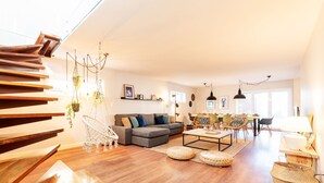Un amplio espacio de sala con una escalera, un conjunto de sofás, una mesa de comedor y plantas colgantes.