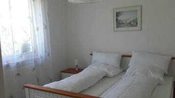 Ein Schlafzimmer mit Bett, Nachttisch, Lampe und Fenster mit durchscheinenden Vorhängen.