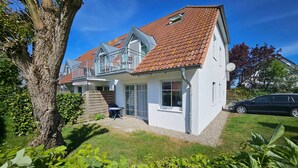 Une maison à deux étages, avec un toit recouvert de tuiles rouges, un balcon et un petit jardin agrémenté de plantes vertes.