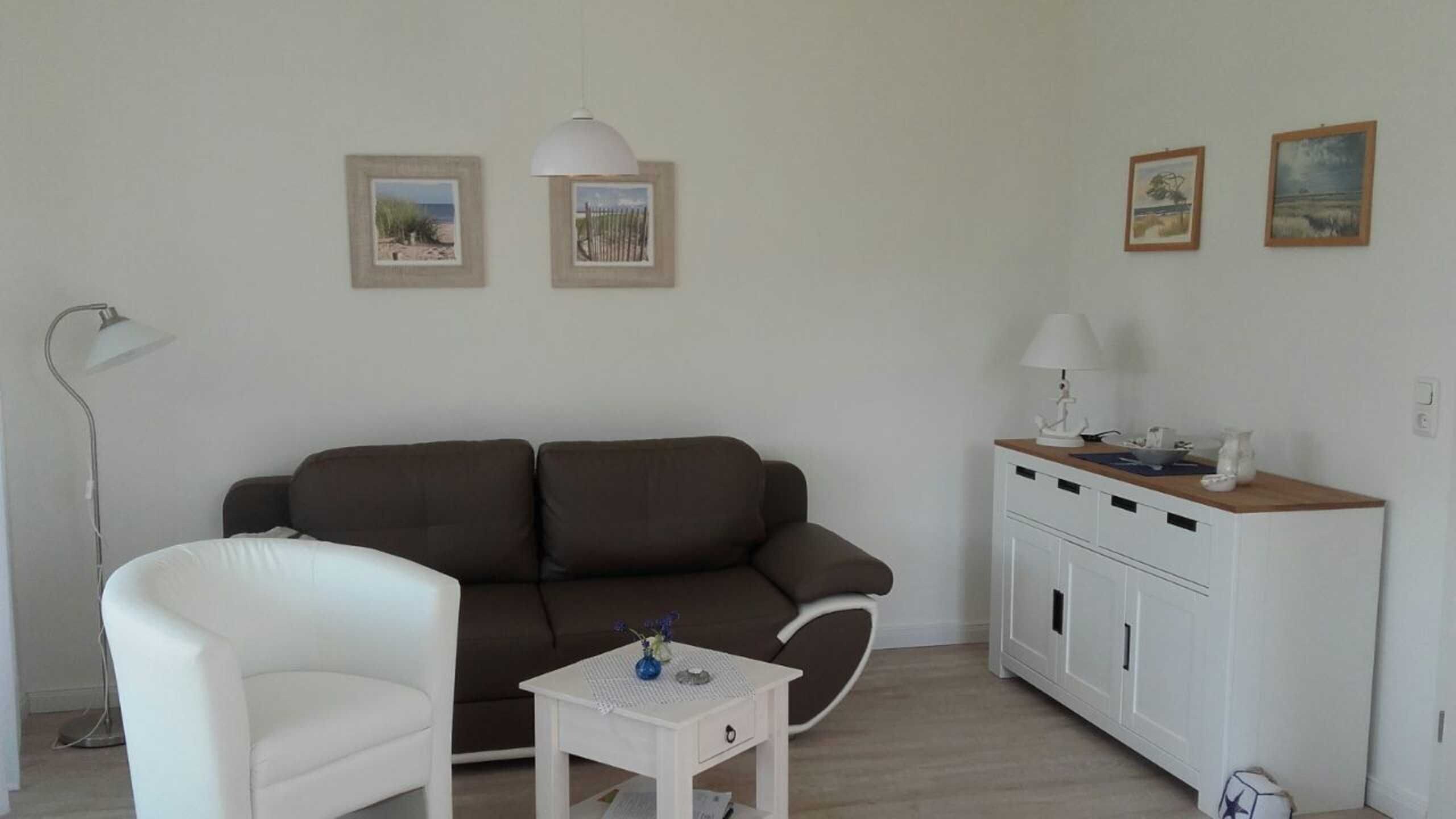 A living room with a brown sofa, a white armchair, a small white table with a vase, and a white cabinet with a lamp on top.
