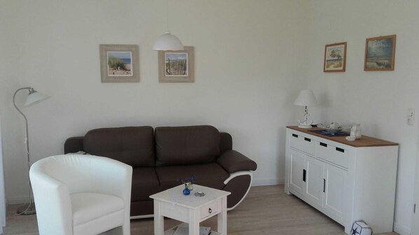Un salon avec un canapé marron, un fauteuil blanc, une petite table blanche ornée d’un vase et un meuble blanc sur lequel est posée une lampe.