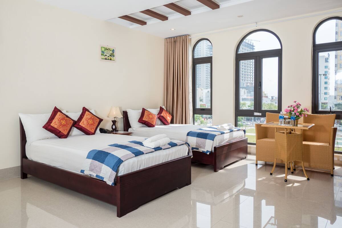 A hotel room with two beds, a small table, and a view of the city through large windows.