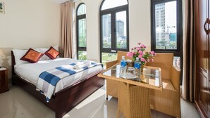 Une chambre d’hôtel avec un lit, une table en verre sur laquelle se trouvent une bouteille et des fleurs, et de grandes fenêtres donnant sur la ville.