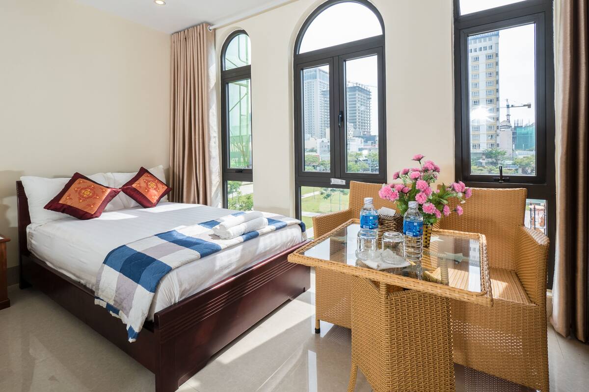 A hotel room with a bed, a glass-top table with a bottle and flowers, and large windows with a city view.