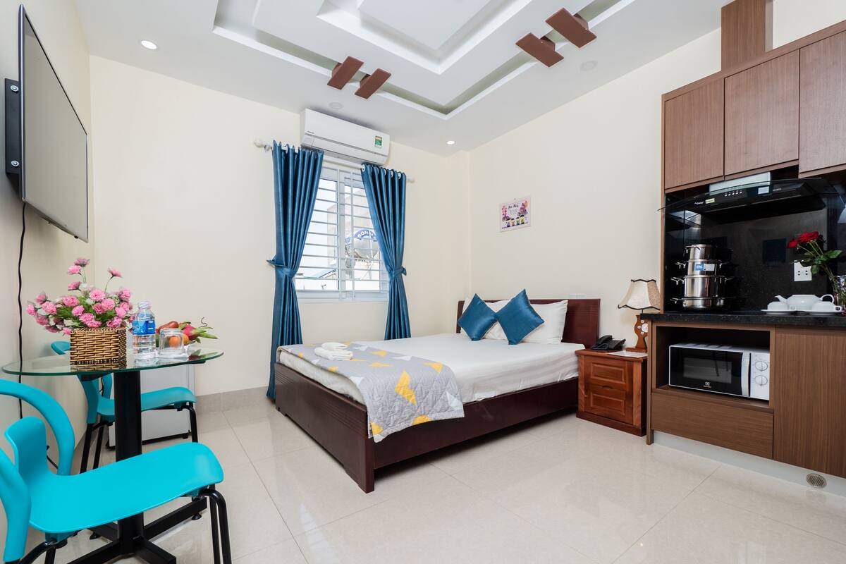 A hotel room with a bed, a desk with a chair, a kitchenette, and a window with curtains.