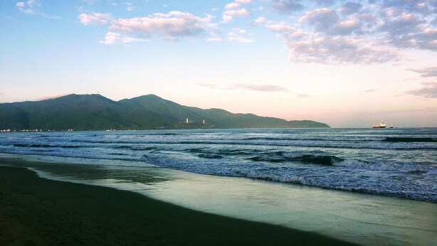 Une plage avec des vagues, une montagne lointaine et un navire sur l’eau.
