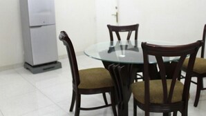 A dining area with a glass table surrounded by wooden chairs with cushioned seats. A white refrigerator is visible in the background. - Furnished Flat In Block C Bashundhara RA (Dhaka)