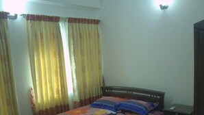 A bedroom with a bed, curtains, a nightstand, and a trash bin. - Furnished Flat In Block C Bashundhara RA (Dhaka)