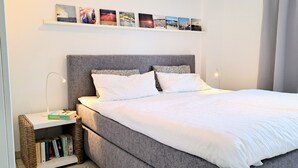 Un lit bien fait, avec du linge de lit blanc, une tête de lit grise et des coussins à motifs. On y voit une bibliothèque remplie de livres et un panier en osier. Au-dessus du lit, une étagère est ornée de cadres avec des photos.