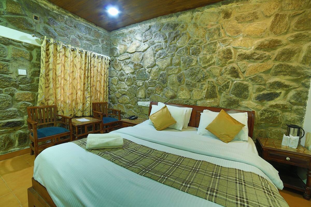 Ein Zimmer mit einer Steinwand, einem Bett mit kariertem Überwurf, zwei Holzstühlen mit blauen Kissen und einem kleinen Tisch.