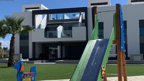 A playground with a green slide and climbing structure, set against a modern building with large windows and a flat roof. - Superb terrace apartment with pool view-Oasis Paradise (Guardamar del Segura)