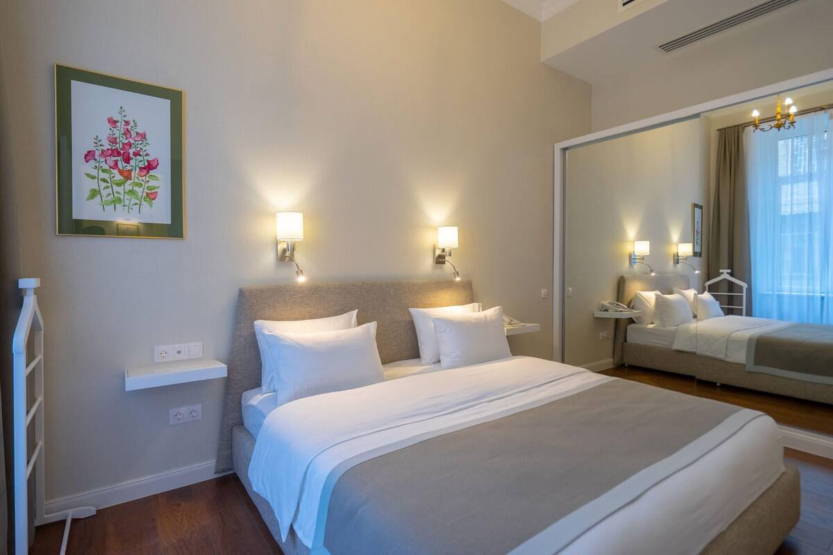 Une chambre d’hôtel avec un grand lit, deux tables de chevet, une lampe fixée au mur, un miroir et un tableau floral encadré.