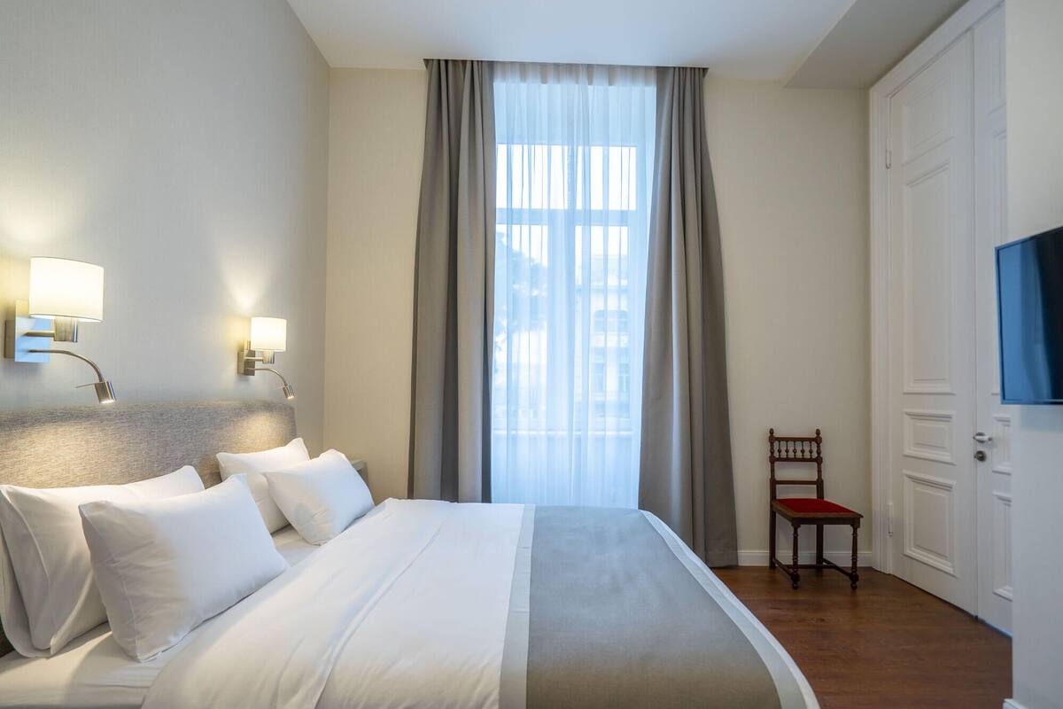 Une chambre d’hôtel avec un lit, une chaise, une télévision et une fenêtre avec des rideaux.