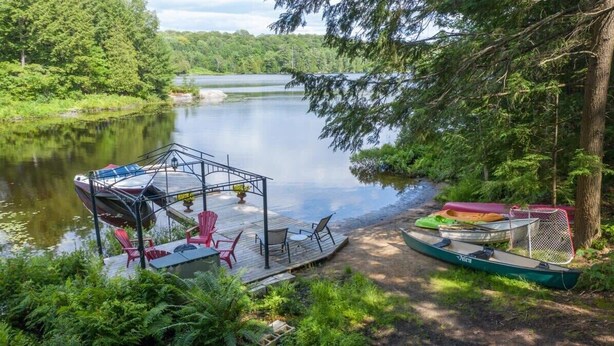 Eine Anlegestelle mit Stühlen und einem Tisch, einem Kanu und einem Boot auf einem stillen See, umgeben von Bäumen.