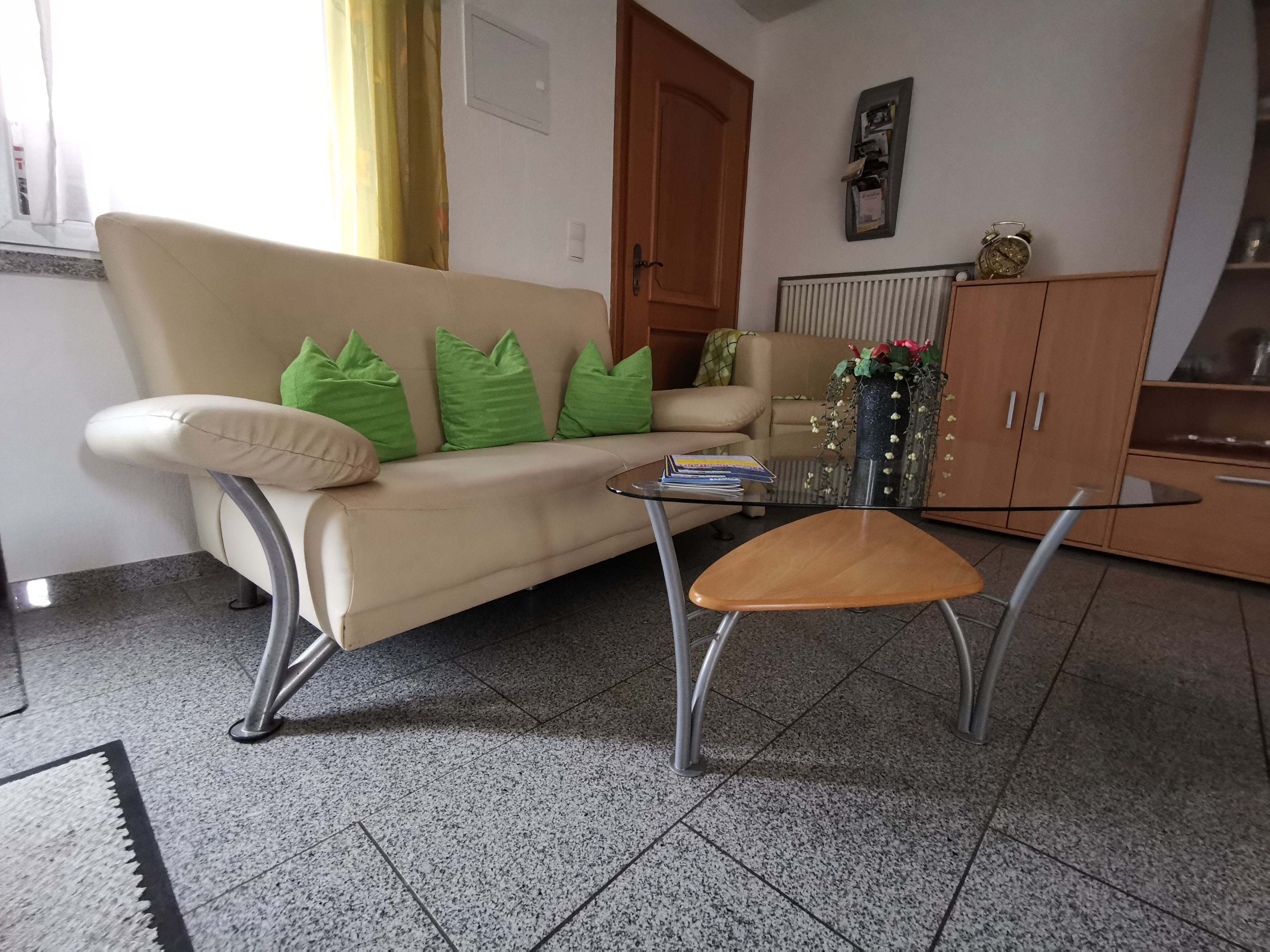 Een woonkamer met een beige bank, groene kussens, een glazen salontafel en een houten kast.