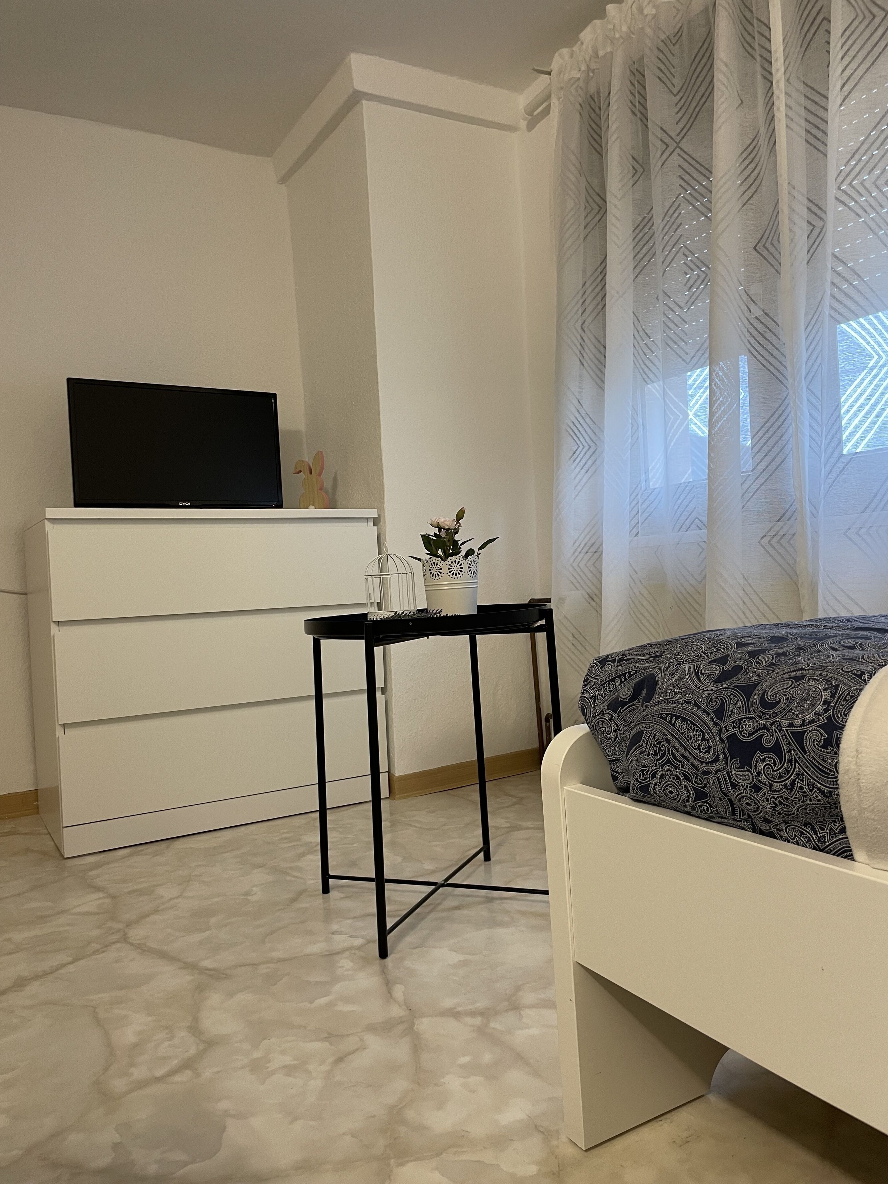 A bedroom with a bed, a TV on a dresser, a small table with a plant, and a window with patterned curtains.