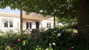 Ein weißes Gebäude mit einem Ziegeldach, einer überdachten Terrasse und einem Garten mit verschiedenen Pflanzen und Blumen.