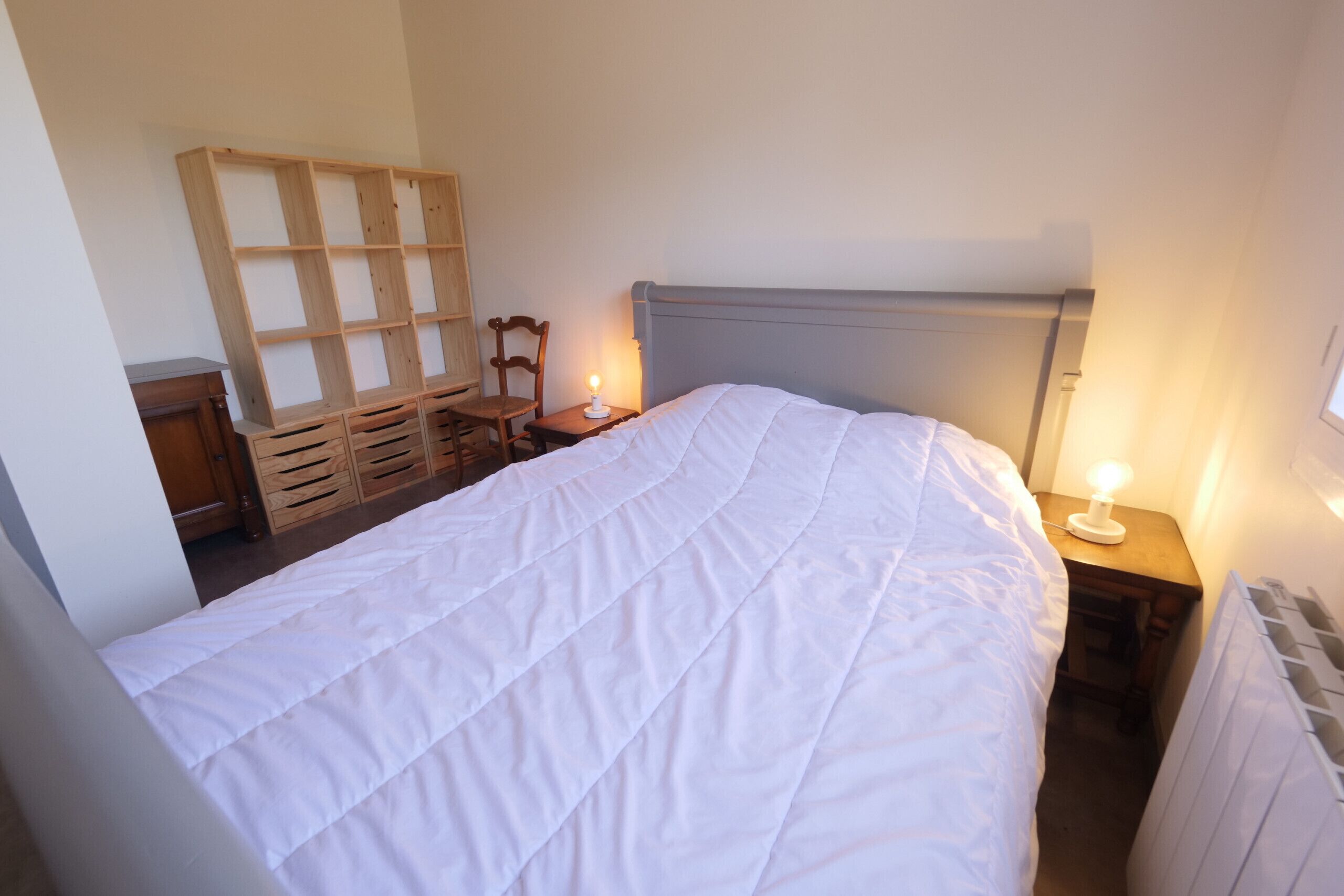 A bedroom with a bed, a nightstand with a lamp, a wooden chair, and a wooden shelf unit.